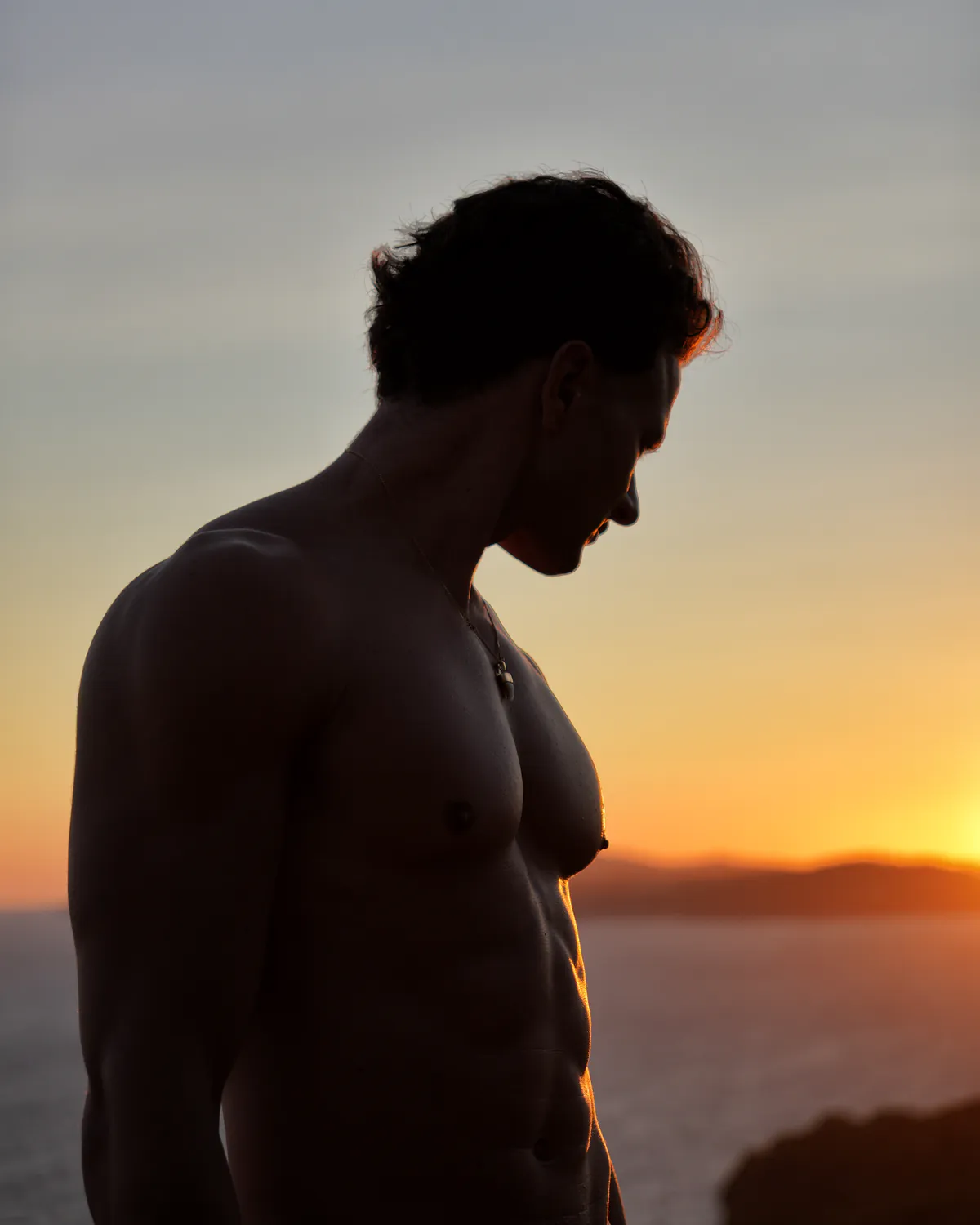 Paul torse nu à contre-jour, la tête baissée, devant la mer au coucher du soleil.