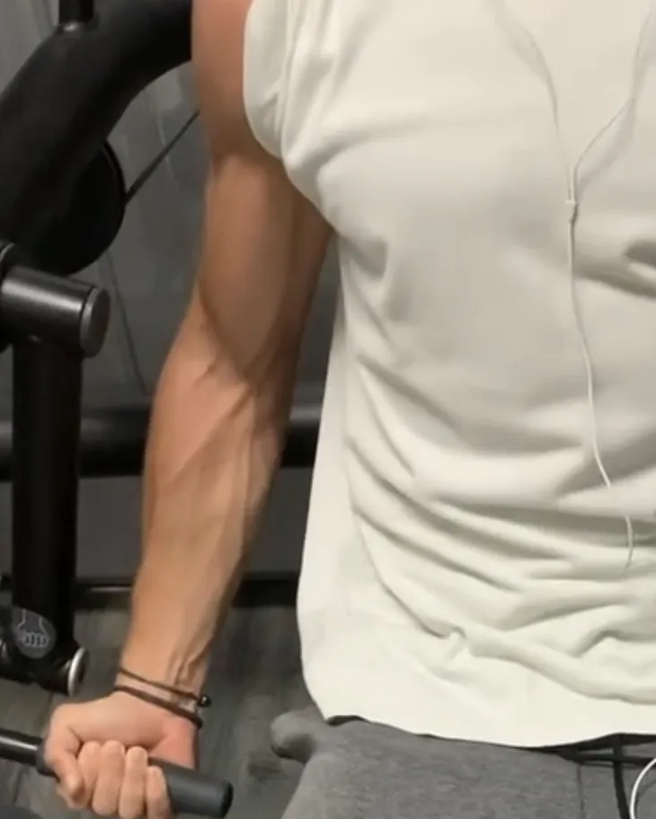 Le bras de Paul, en débardeur blanc, qui tient la poignée d'une machine de musculation, le visage hors du cadre.